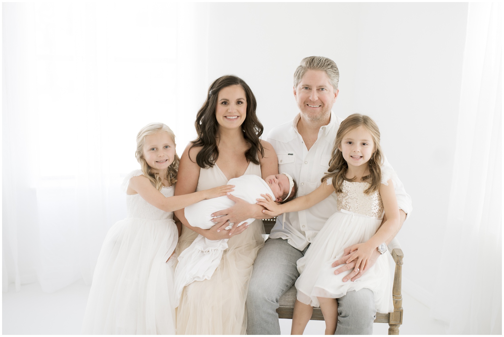 Happy mom and dad sit on a bench in a studio with their newborn daughter and 2 toddler daughters