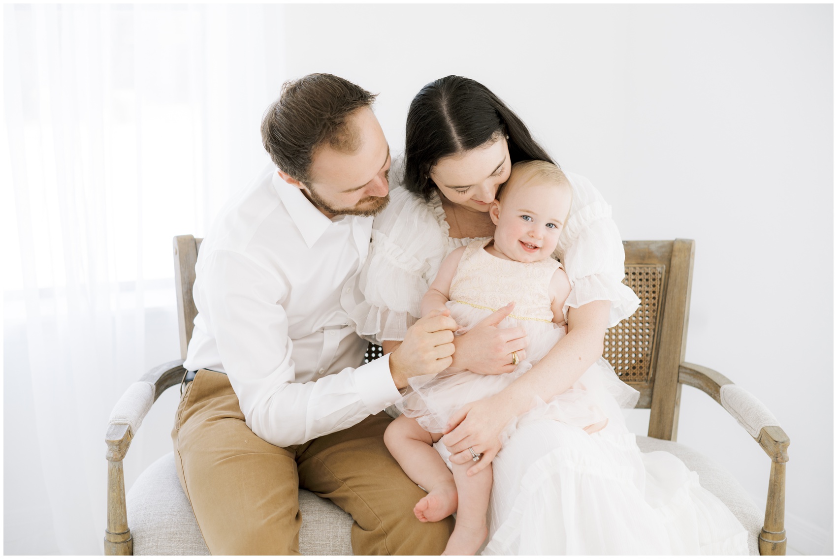 A toddler girl in a pink dress smilies while sitting in mom and dad's laps on a wooden bench