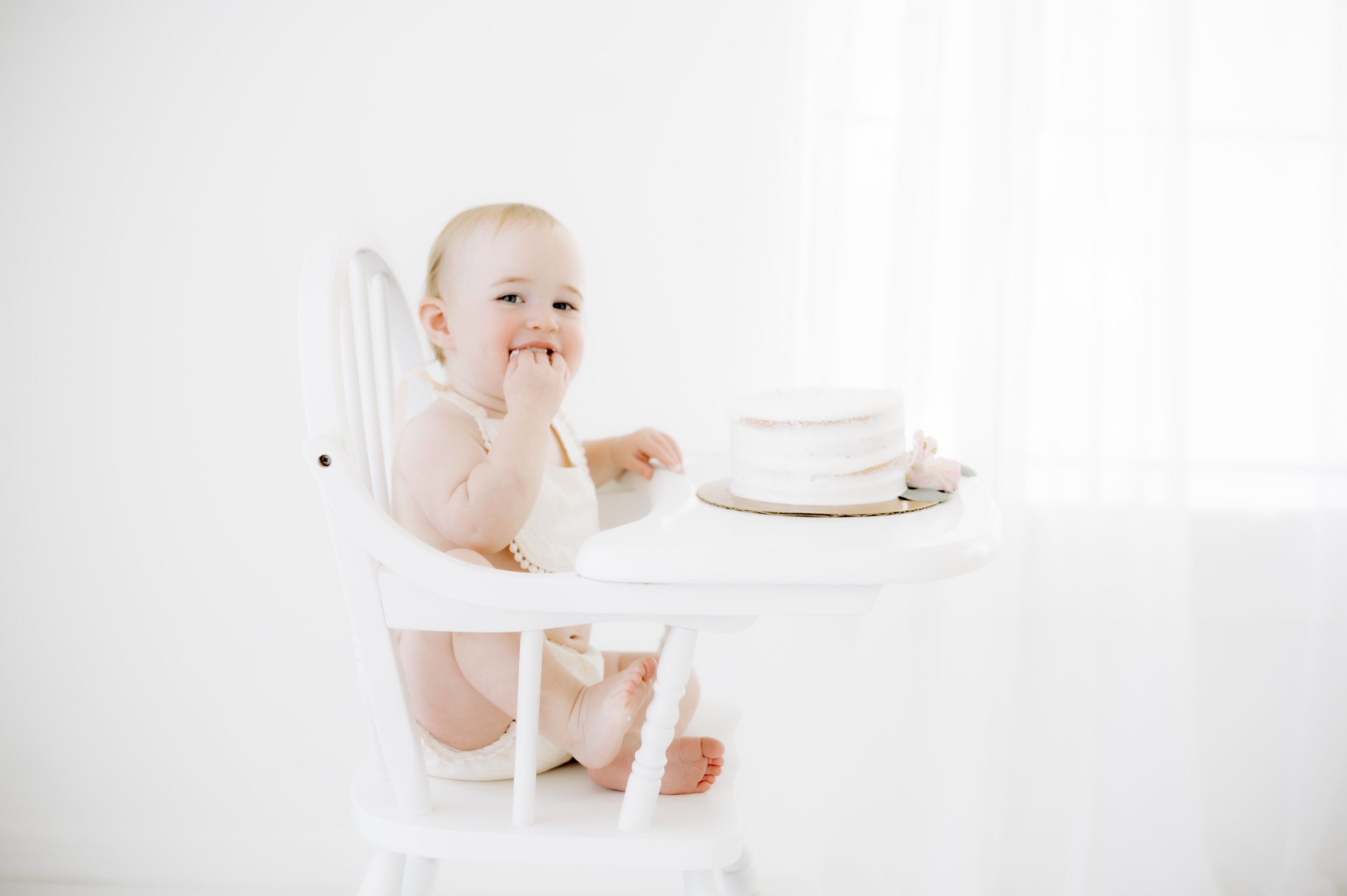 A smiling baby girl puts cake in her mouth while sitting in a white wooden high chair in a studio in a bib