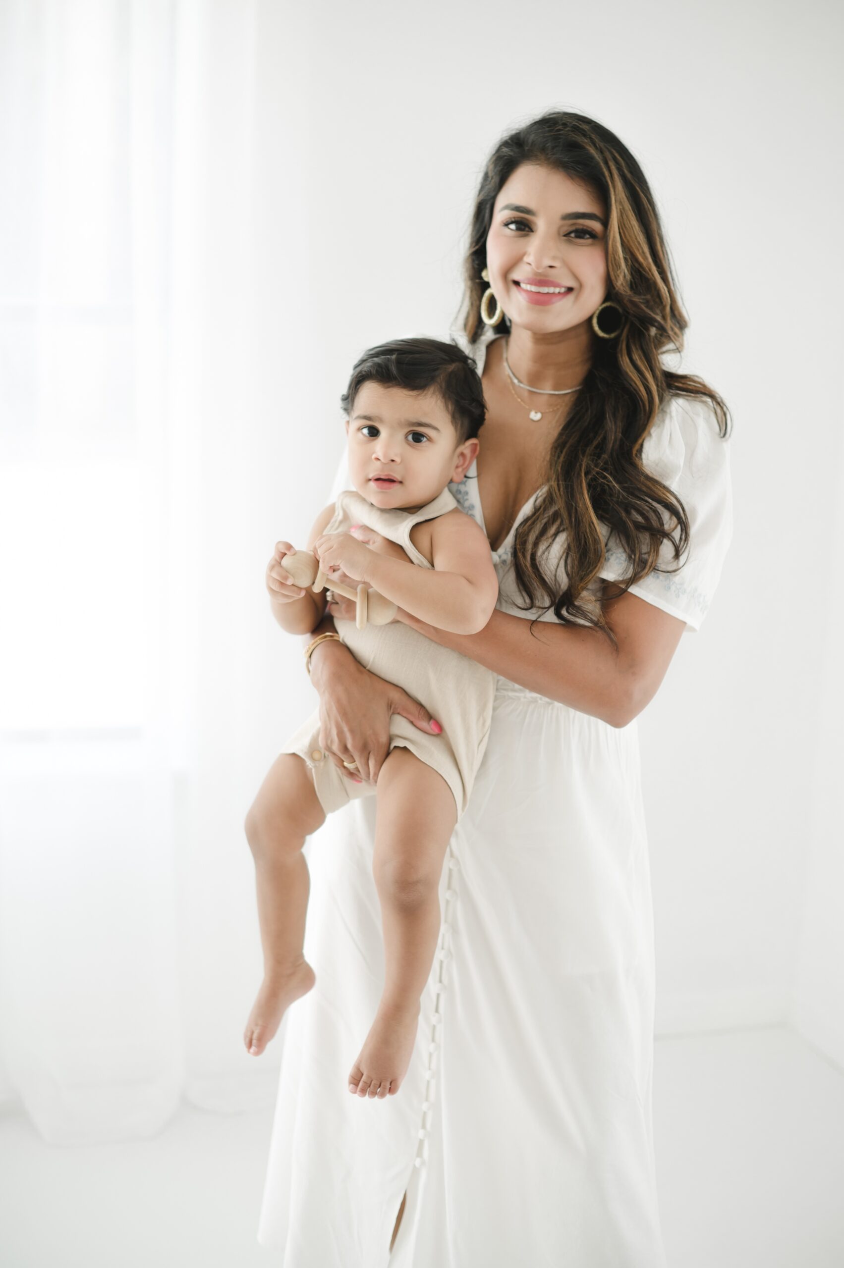 A baby boy in tan overalls sits in mom's arms while holding a wooden toy in a white studio