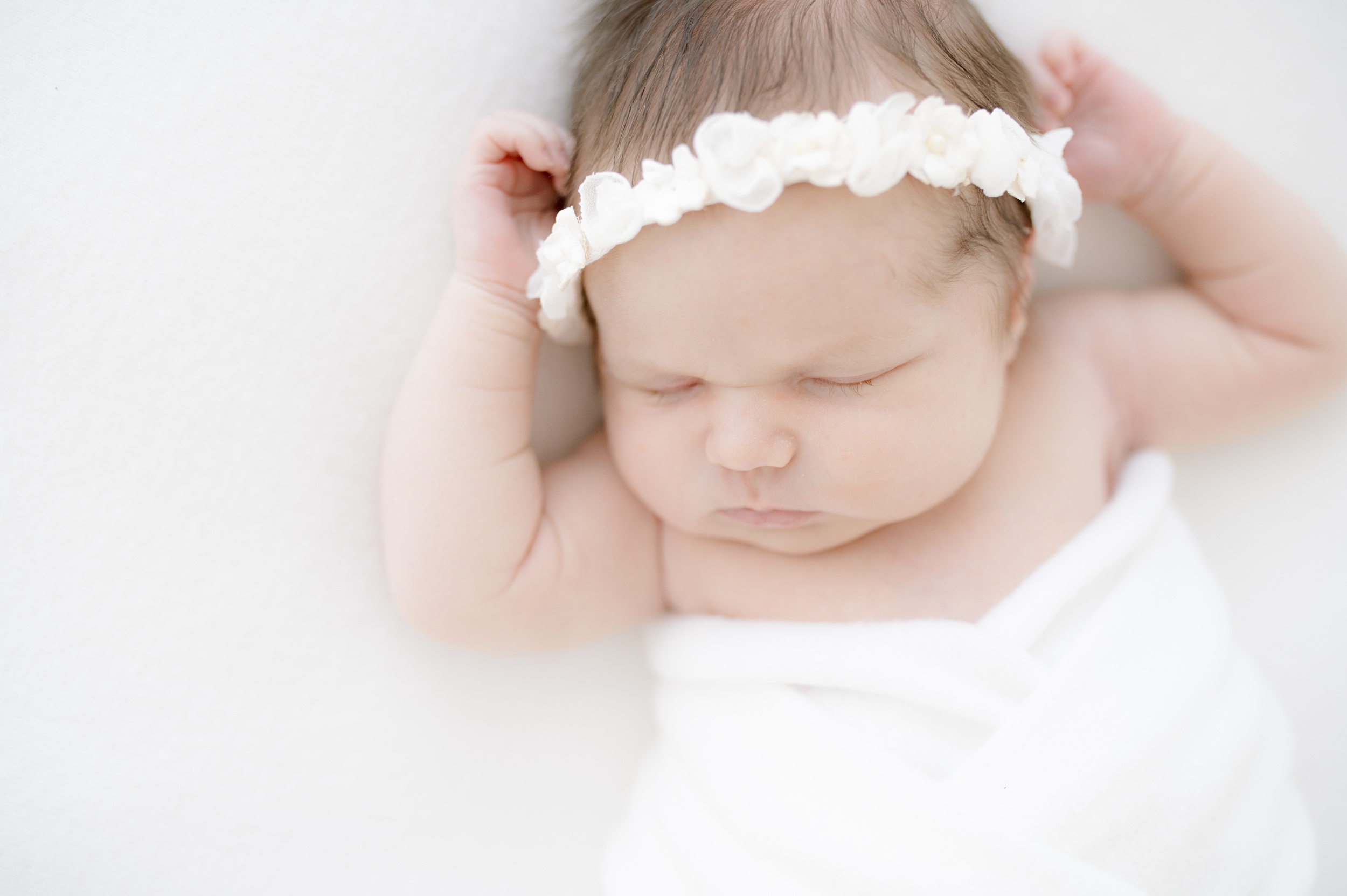 A newborn baby sleeps with arms up in a white blanket and flower headband