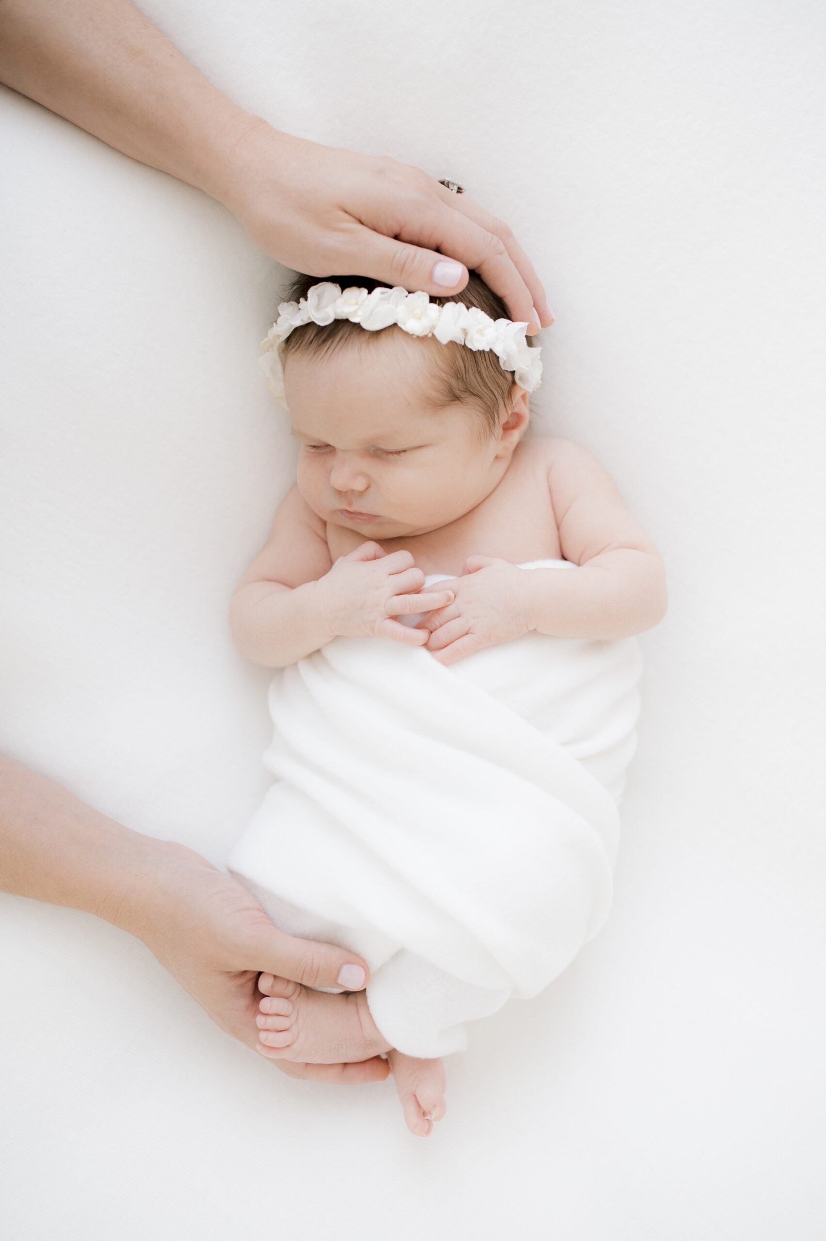 A newborn baby girl sleeps in a white swaddle and flower headband with mom's hands on feet and head before some infant swim lessons in austin