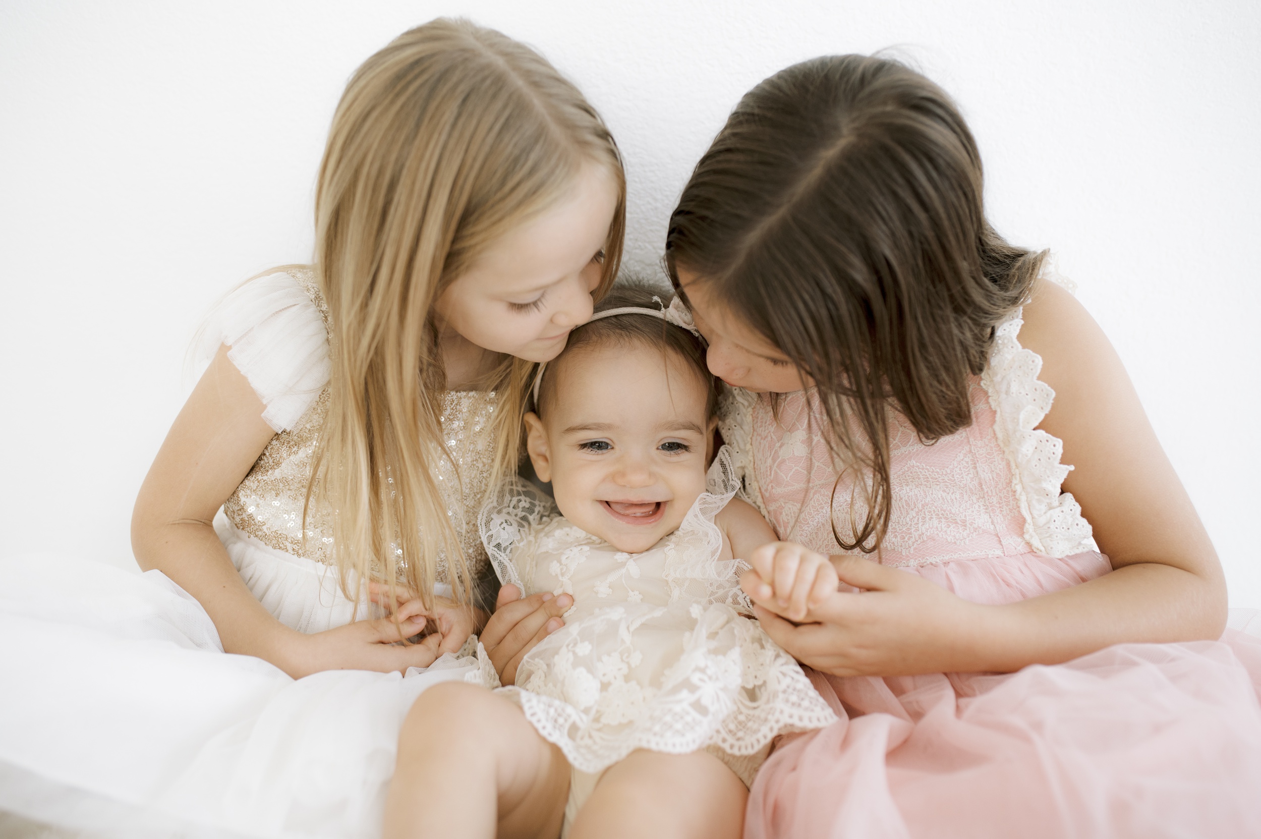 A smiling infant girl is kissed and hugged by her bigger toddler sisters while sitting in white and pink dresses