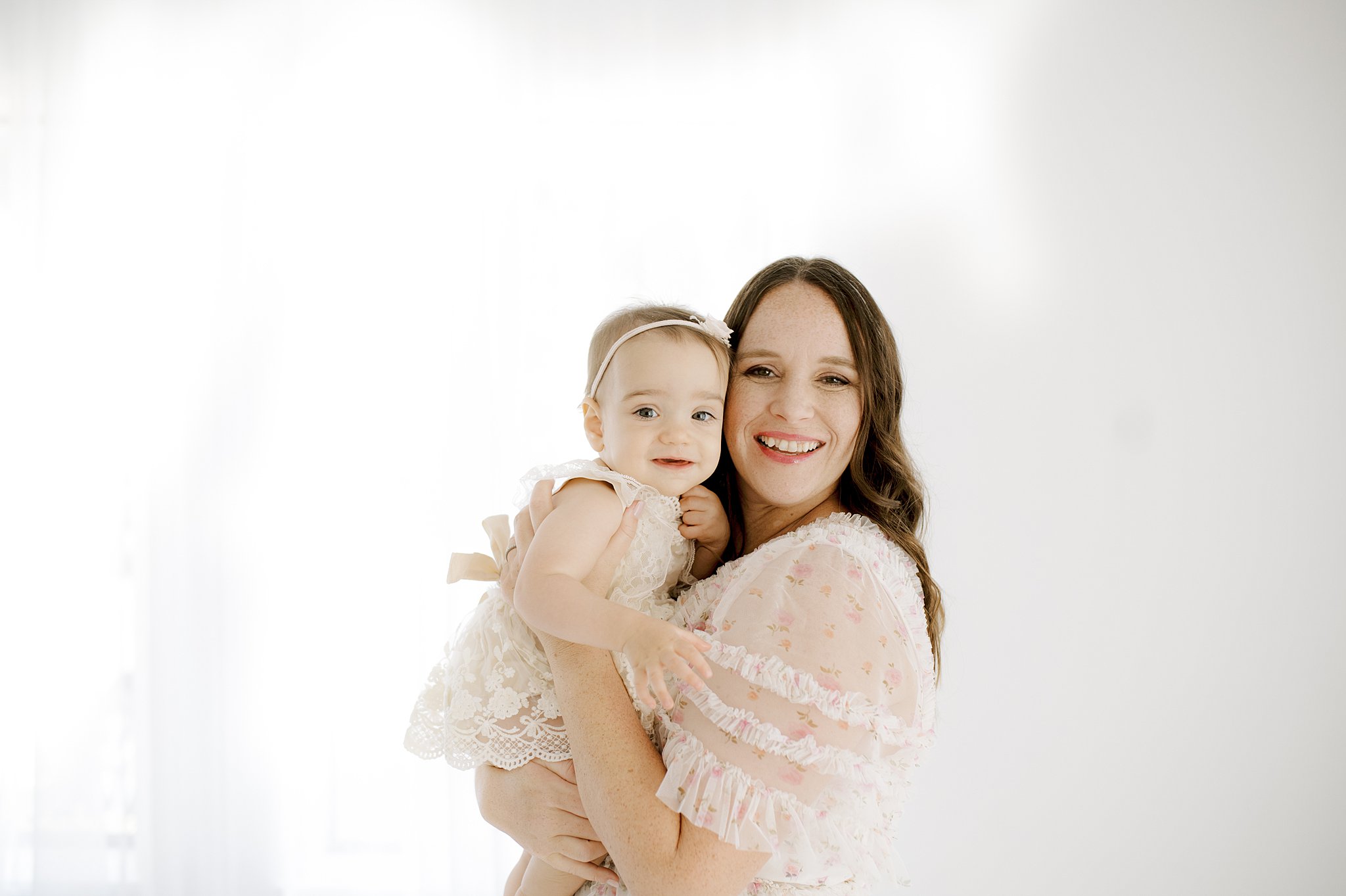 A happy mom stands in a studio smiling with her baby daughter in her arms against her cheek in white lace dresses after exploring new year's eve in austin with kids