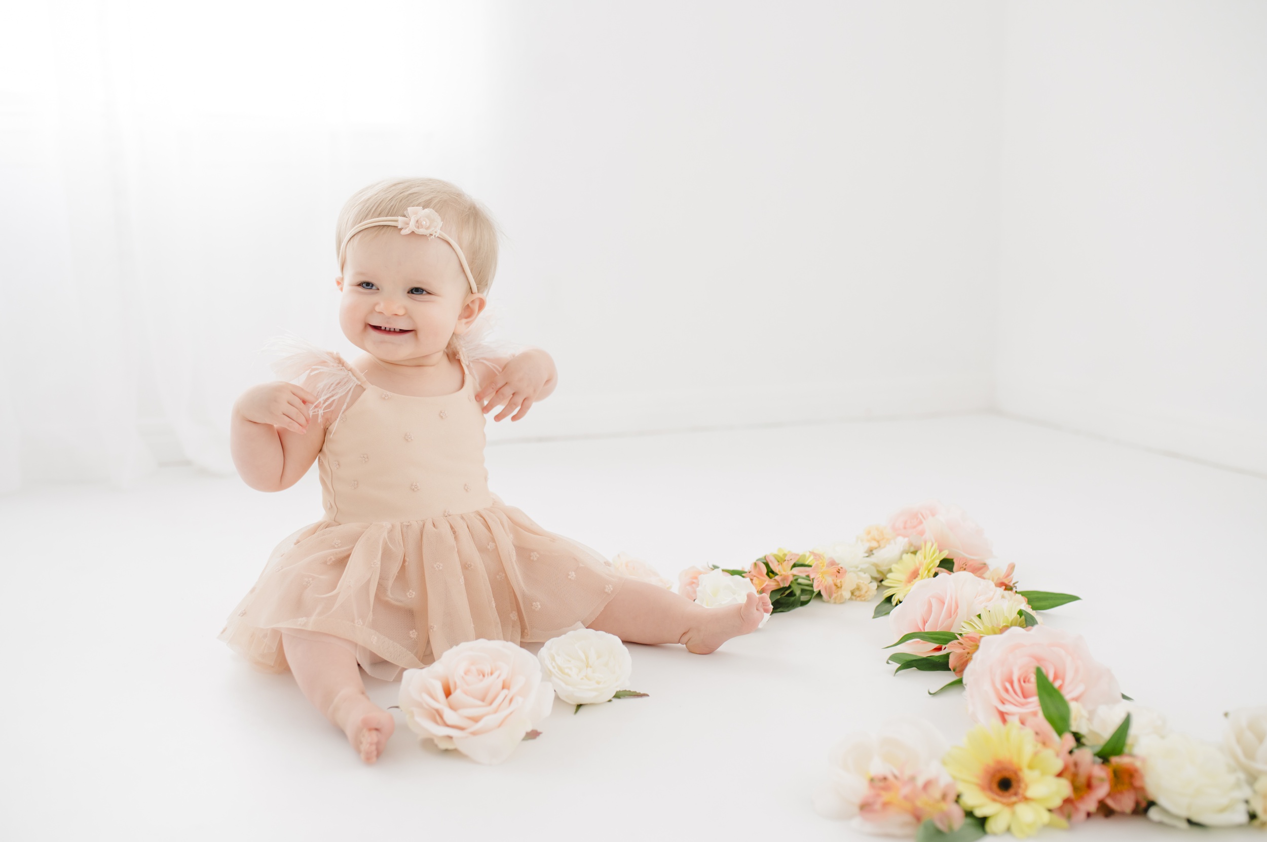 A smiling baby sits in a cream dress surrounded by flowers in a studio after visiting preschools in austin